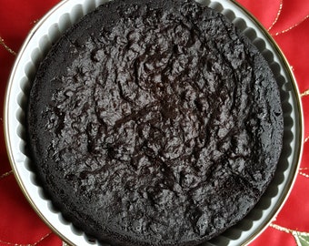 May include: A dark chocolate cake in a white baking pan with a fluted edge. The cake is round and has a slightly cracked surface.