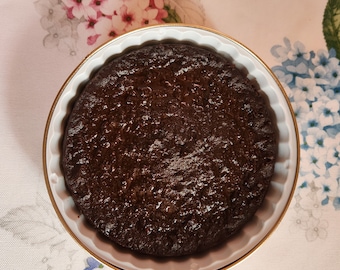 May include: A round chocolate cake in a white baking dish with a gold rim. The cake is dark brown and has a shiny, glazed surface.