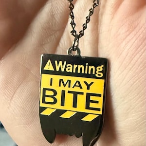 Może przedstawiać: Naszyjnik z czarno-żółtą zawieszką z napisem "A Warning I May Bite". Zawieszka ma kształt kła i wisi na brązowym łańcuszku.