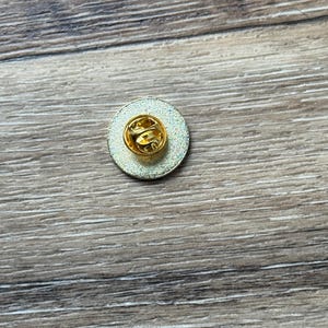 May include: A circular metal pin badge shown from the reverse side, featuring a gold-toned butterfly clutch fastener centered on a textured, metallic silver-colored disc. The pin mechanism is clearly visible, showcasing the standard hardware.