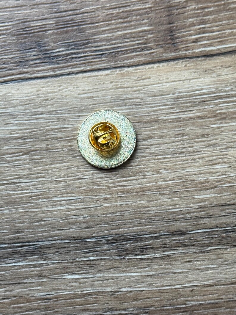 May include: A circular metal pin badge shown from the reverse side, featuring a gold-toned butterfly clutch fastener centered on a textured, metallic silver-colored disc. The pin mechanism is clearly visible, showcasing the standard hardware.