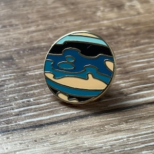 May include: A circular enamel pin featuring a stylized planet design with a gold-toned metal border. The surface displays wavy, abstract bands of color in shades of black, cream, light blue, and dark teal, creating a layered, topographic appearance.