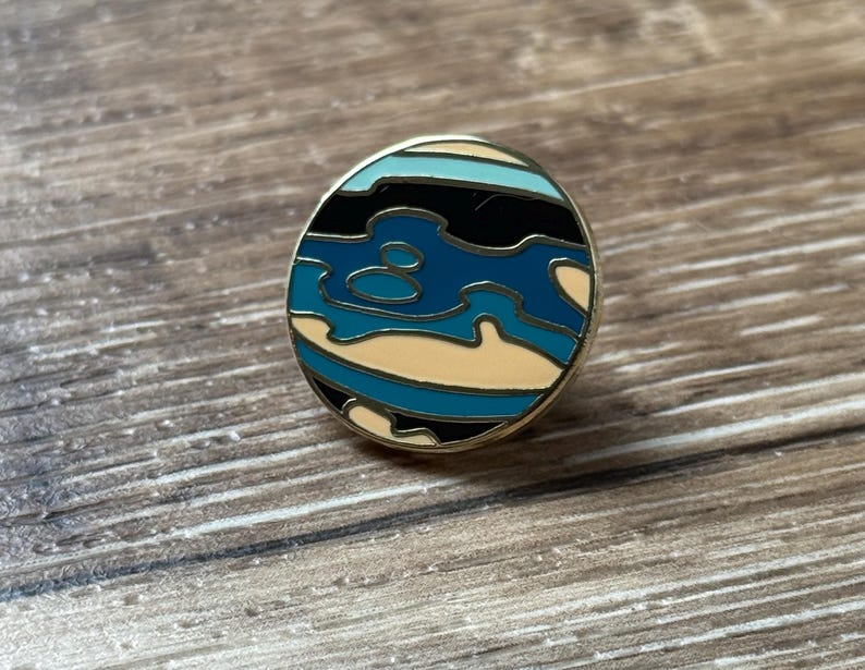 May include: A circular enamel pin featuring a stylized planet design with a gold-toned metal border. The surface displays wavy, abstract bands of color in shades of black, cream, light blue, and dark teal, creating a layered, topographic appearance.