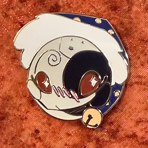 May include: Enamel pin featuring a stylized crescent moon and a sleeping figure. The pin has a white, black, and cream color scheme with accents of red, pink, and gold. A small gold bell hangs at the bottom.