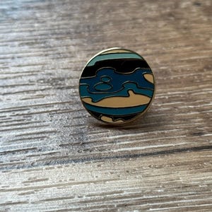 May include: A circular enamel pin featuring a stylized abstract design with horizontal bands of black, light blue, dark blue, and cream colors. The pin has a thin gold-toned metal border that frames the colorful enamel inlay in a round shape.