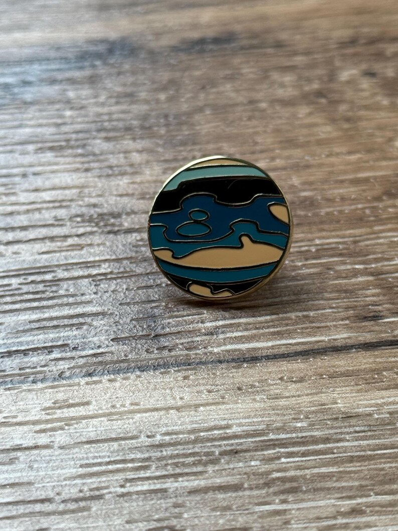 May include: A circular enamel pin featuring a stylized abstract design with horizontal bands of black, light blue, dark blue, and cream colors. The pin has a thin gold-toned metal border that frames the colorful enamel inlay in a round shape.