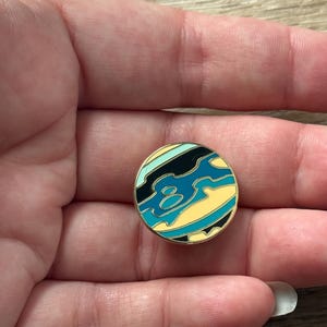 May include: A circular enamel pin featuring a stylized abstract design with wavy bands of black, teal, light blue, and pale yellow. The pin has a thin gold-toned metal border that outlines the colorful sections, creating a smooth, glossy surface.