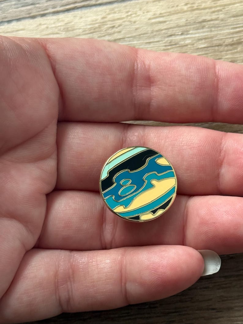 May include: A circular enamel pin featuring a stylized abstract design with wavy bands of black, teal, light blue, and pale yellow. The pin has a thin gold-toned metal border that outlines the colorful sections, creating a smooth, glossy surface.