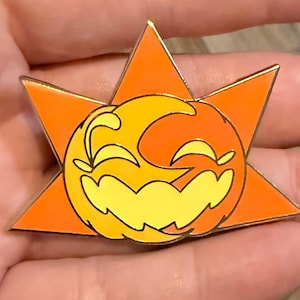 May include: A gold-edged enamel pin featuring a smiling sun-moon hybrid design. The pin has a bright orange starburst shape with a yellow and orange smiling face in the center. A fun accessory for bags, jackets, or hats.
