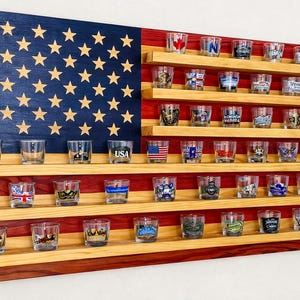 May include: Wooden shot glass display in the shape of the American flag. The blue section has gold stars, and the red and white stripes hold multiple shot glasses. Each shot glass has a unique design.