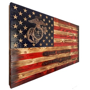 May include: A wooden American flag with a distressed, burnt look. The flag features the Marine Corps emblem and the text "SGT GUILLERMO CAMACHO UNITED STATES MARINE CORPS".