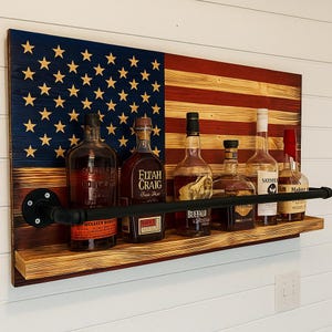 May include: A wooden wall-mounted bar shelf with an American flag design. The shelf holds several liquor bottles and features a black metal pipe rail. The flag design is made of red, white, and blue wood.