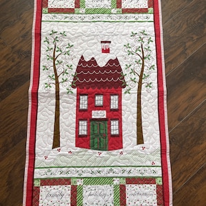May include: A red and green quilted wall hanging with a white background. The center of the hanging features a red house with a green door and white snow on the roof. The house is surrounded by two trees with green leaves and red berries. The top of the hanging has the words "peace", "love", and "joy" in red squares. The bottom of the hanging has the words "family", "holidays", and "celebration" in red and green squares.