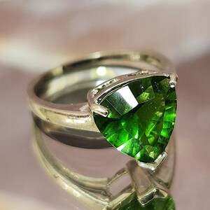 May include: A silver ring with a large, triangular, green gemstone. The ring is set on a white, textured surface.