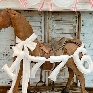 May include: A rustic rocking horse made of burlap with a leather saddle and white fabric accents. The number "1776" is attached to the horse. A patriotic garland with red, white, and blue bows is above the horse.