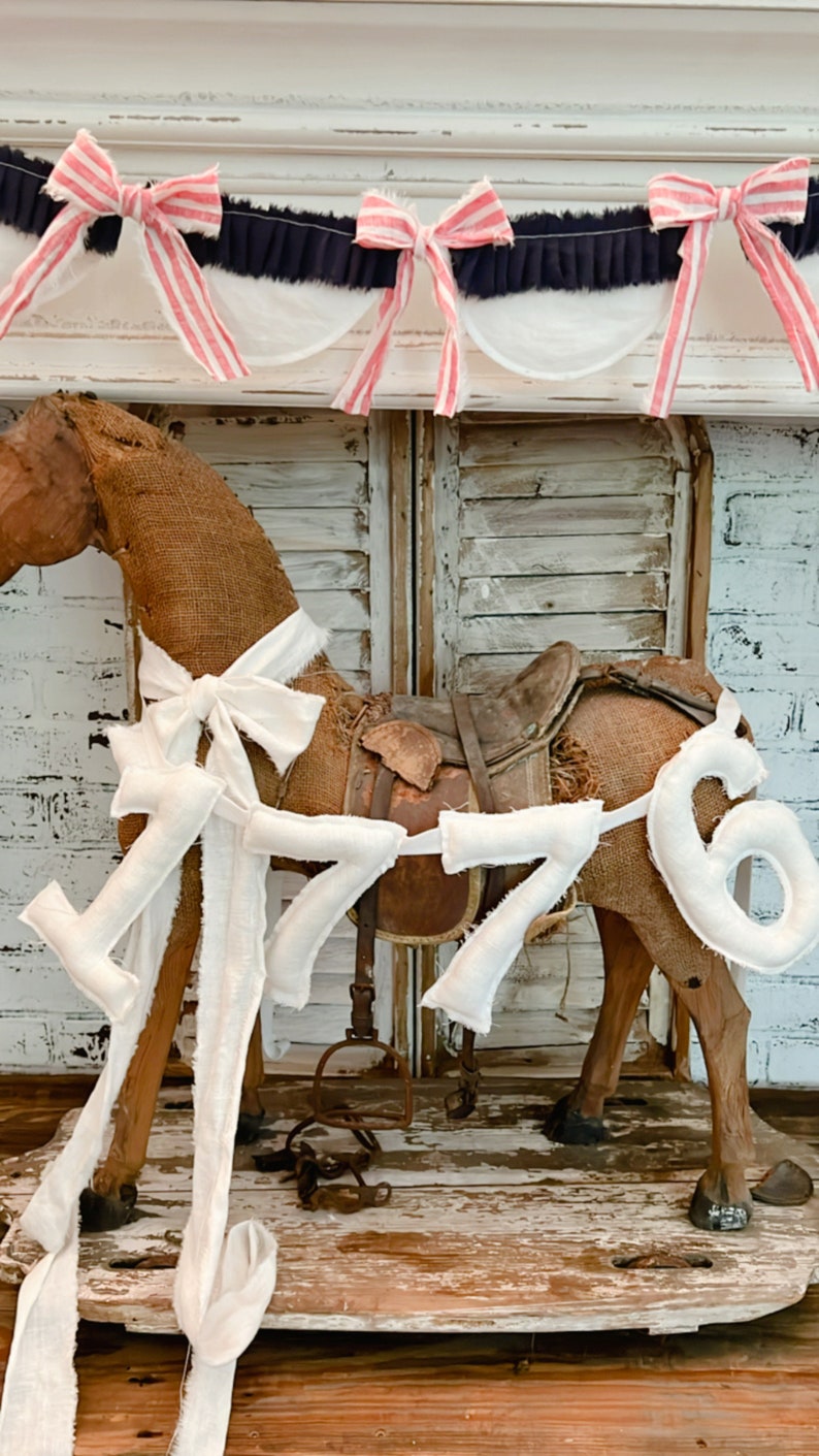 May include: A rustic rocking horse made of burlap with a leather saddle and white fabric accents. The number "1776" is attached to the horse. A patriotic garland with red, white, and blue bows is above the horse.