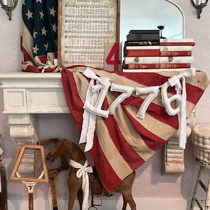 May include: A patriotic display featuring an American flag, a framed print with sheet music, and a banner reading "1776." The scene includes a wooden rocking horse, a vintage tennis racket, and a stack of books, all arranged on a white mantel.
