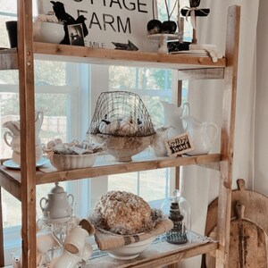 May include: A rustic wooden shelf unit with three shelves, decorated with white farmhouse style decor, including a sign that reads "Pumpkin Cottage Farm 1 Mile", a birdcage, and a white pumpkin.