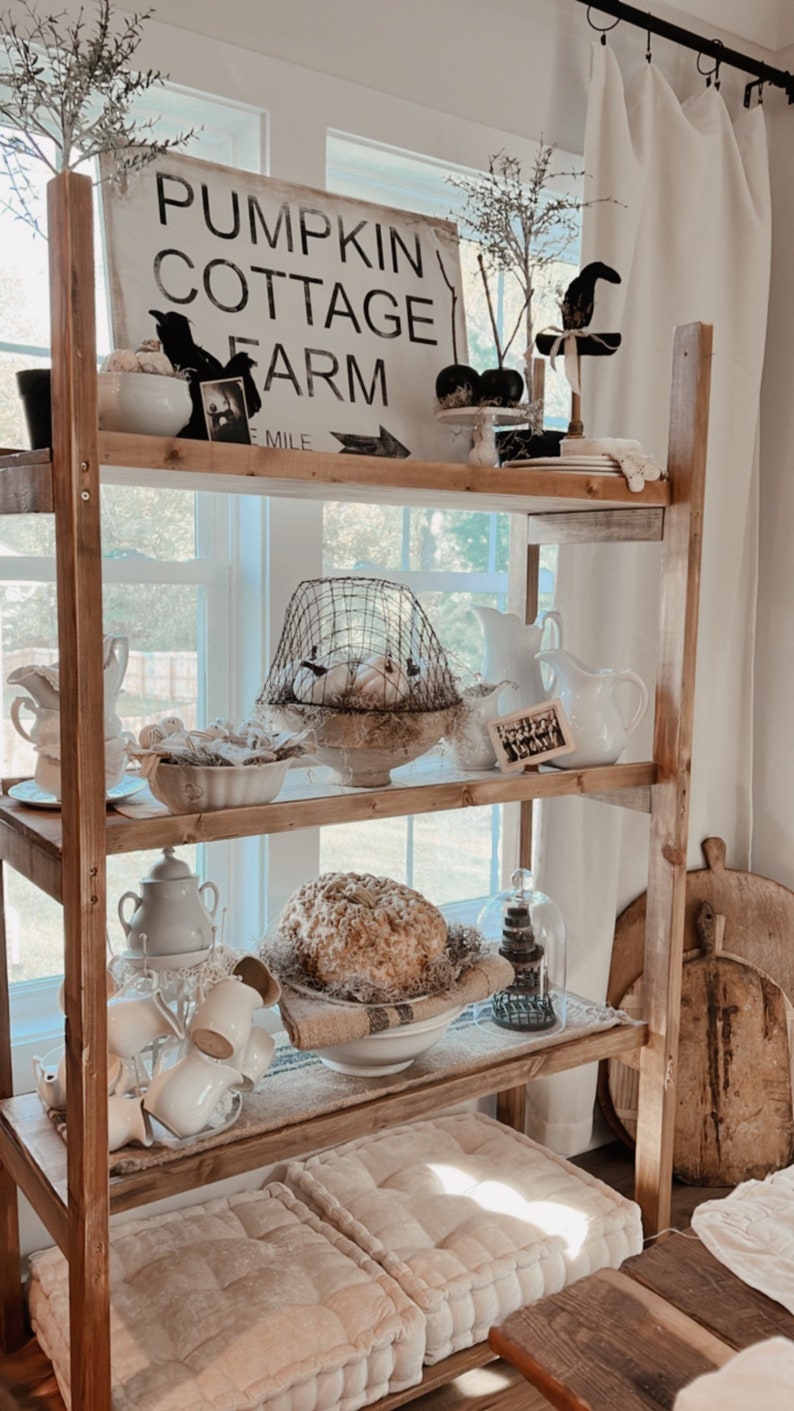 May include: A rustic wooden shelf unit with three shelves, decorated with white farmhouse style decor, including a sign that reads "Pumpkin Cottage Farm 1 Mile", a birdcage, and a white pumpkin.