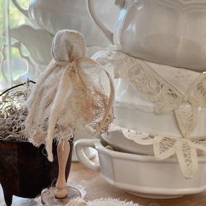 May include: A white fabric doll with a white fabric dress and a wooden base. The doll is sitting on a wooden surface with a white fabric banner that says "boo" on it. There are white ceramic bowls and lace doilies in the background.