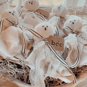 May include: A white bowl filled with white fabric ghosts with black eyes and mouths. Each ghost has a small brown tag with the word "boo" written on it. The ghosts are sitting on a bed of brown dried moss.