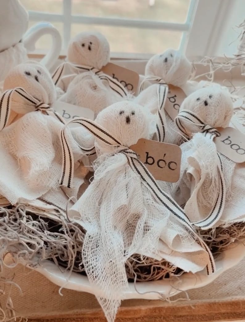 May include: A white bowl filled with white fabric ghosts with black eyes and mouths. Each ghost has a small brown tag with the word "boo" written on it. The ghosts are sitting on a bed of brown dried moss.