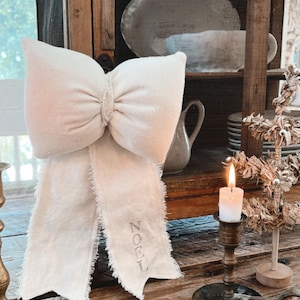 May include: A white fabric bow with frayed edges and the word "NOEL" embroidered on it. The bow is displayed on a wooden table with a candle and a small artificial Christmas tree.