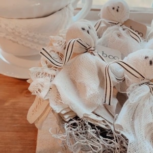 May include: A white bowl filled with shredded white paper and four white fabric ghosts with black ribbon tied around their necks. The ghosts have black eyes and mouths drawn on them. The bowl is sitting on a wooden surface with a white lace doily underneath.