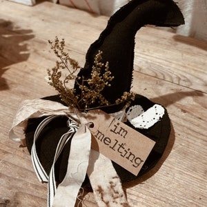 May include: A black witch hat with a brown tag that says "I'm melting". The hat is decorated with a white lace trim and a sprig of dried flowers.