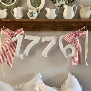 May include: A display of vintage plates and cream pitchers on a wooden shelf. Below, a banner spelling "1776" with red and white striped bows hangs above white pillows, creating a patriotic and farmhouse aesthetic.