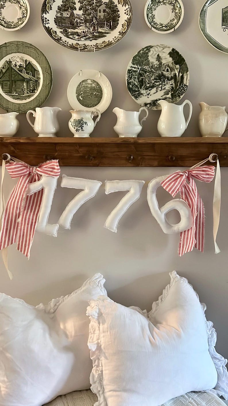 May include: A display of vintage plates and cream pitchers on a wooden shelf. Below, a banner spelling "1776" with red and white striped bows hangs above white pillows, creating a patriotic and farmhouse aesthetic.