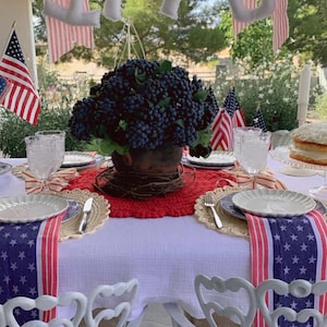 May include: A festive outdoor table setting with patriotic decor. Features a white tablecloth, star-spangled runners, and decorative plates. A centerpiece of dark blue berries sits in a rustic pot. The number "1776" hangs above the table.