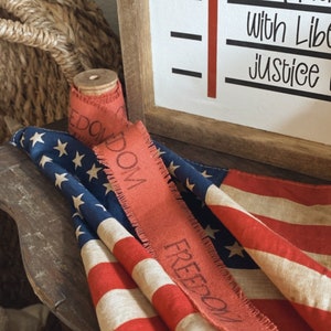 May include: A red, white, and blue American flag with a red ribbon that says "FREEDOM DOWN" on it. The flag is folded and draped over a wooden surface.