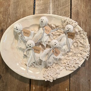 May include: Six white fabric ghosts with black and white ribbon tails on a white plate with a crocheted doily. Each ghost has a brown tag with the word "boo" written on it.