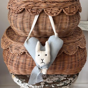 May include: Two brown wicker baskets with scalloped edges, stacked on top of each other. A blue heart-shaped fabric toy with a white bunny face and a blue scarf hangs from the top basket.