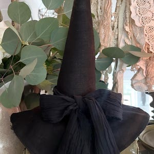 May include: A black witch hat with a tall, pointed crown and a wide brim. A large, black fabric bow is tied around the base of the crown. The hat is likely made of fabric and is a decorative item.