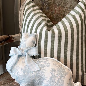May include: A blue and white toile patterned bunny rabbit pillow with a blue and white gingham bow. The bunny is sitting on a chair with a green and white striped pillow.