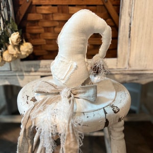 May include: A white linen witch hat with a frayed edge and a brown ribbon bow. The hat is sitting on a white stool.