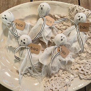 May include: Five white gauze ghosts with black eyes and mouths, each with a brown tag that says "boo" or "boo" on a white plate with a lace doily.