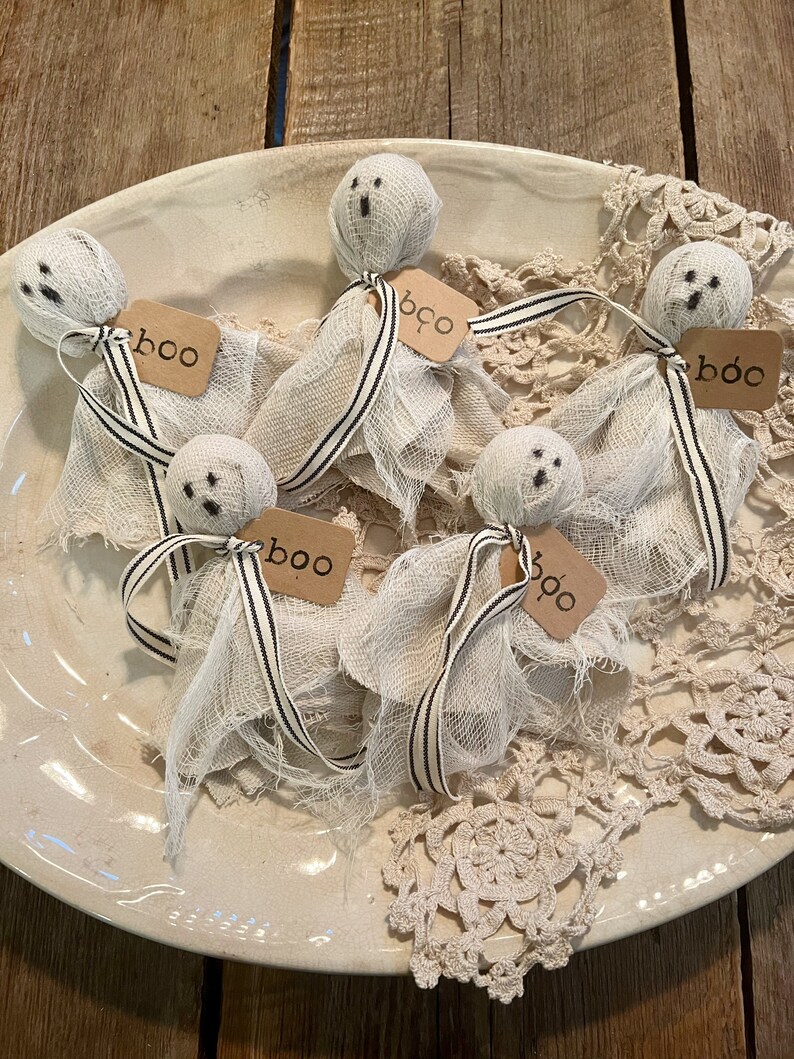 May include: Five white gauze ghosts with black eyes and mouths, each with a brown tag that says "boo" or "boo" on a white plate with a lace doily.