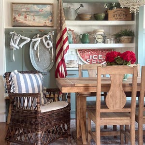 May include: A dining room scene with a wicker armchair, wooden table, and chairs. A vintage American flag hangs in the background. Shelves display decorative items, including a sign that reads "ICE COLD". A painting and a chandelier are also visible.
