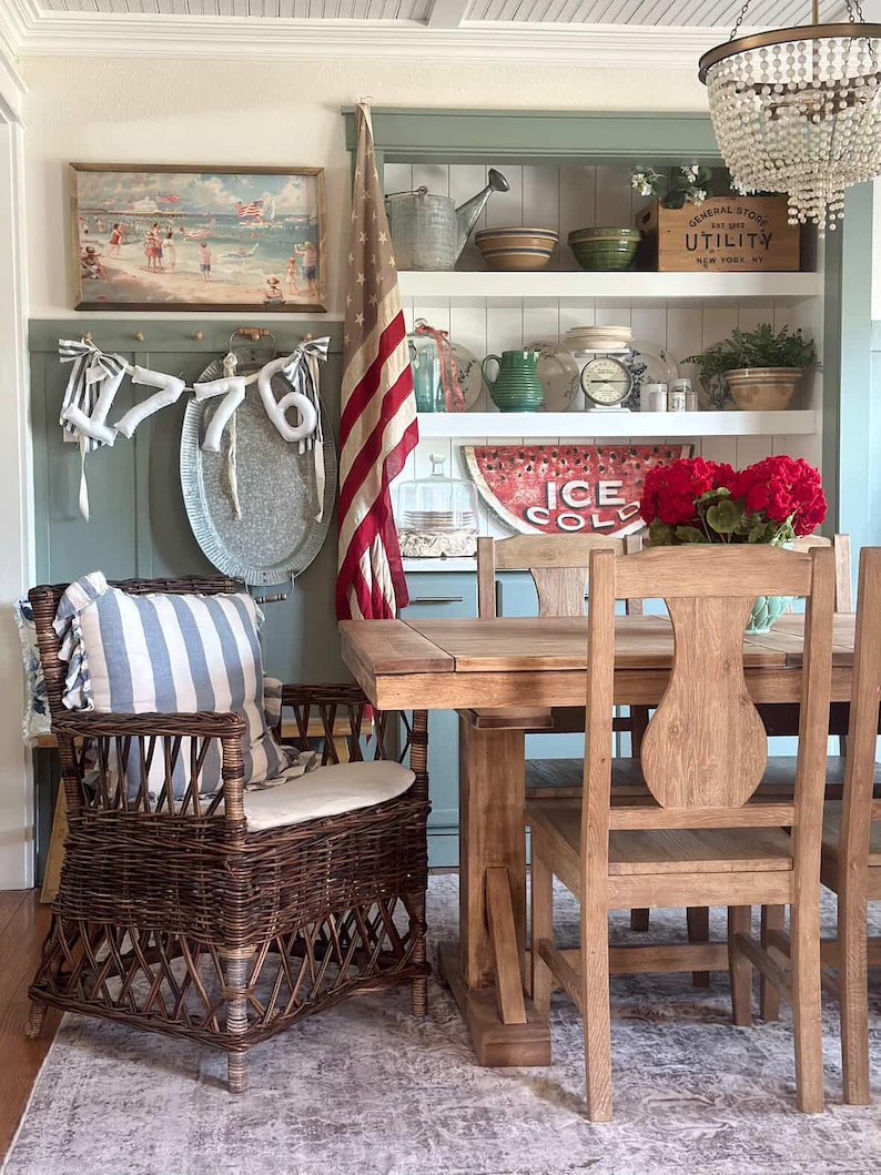May include: A dining room scene with a wicker armchair, wooden table, and chairs. A vintage American flag hangs in the background. Shelves display decorative items, including a sign that reads "ICE COLD". A painting and a chandelier are also visible.