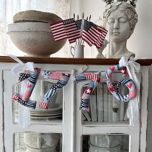 May include: A patriotic 1776 banner with red, white, and blue fabric, adorned with stars and hearts. The banner is displayed on a white cabinet, complemented by American flags and a bust sculpture, creating a festive, decorative theme.
