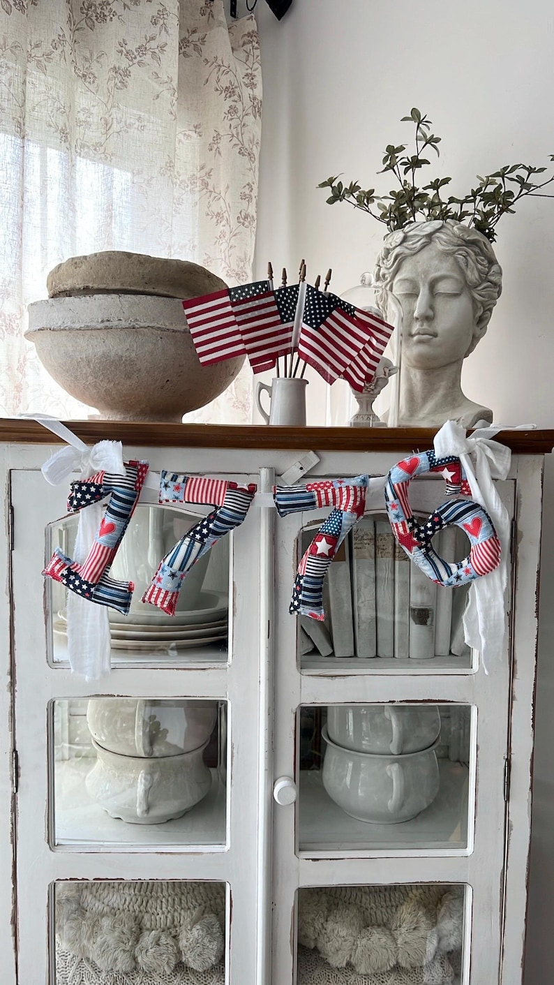 May include: A patriotic 1776 banner with red, white, and blue fabric, adorned with stars and hearts. The banner is displayed on a white cabinet, complemented by American flags and a bust sculpture, creating a festive, decorative theme.