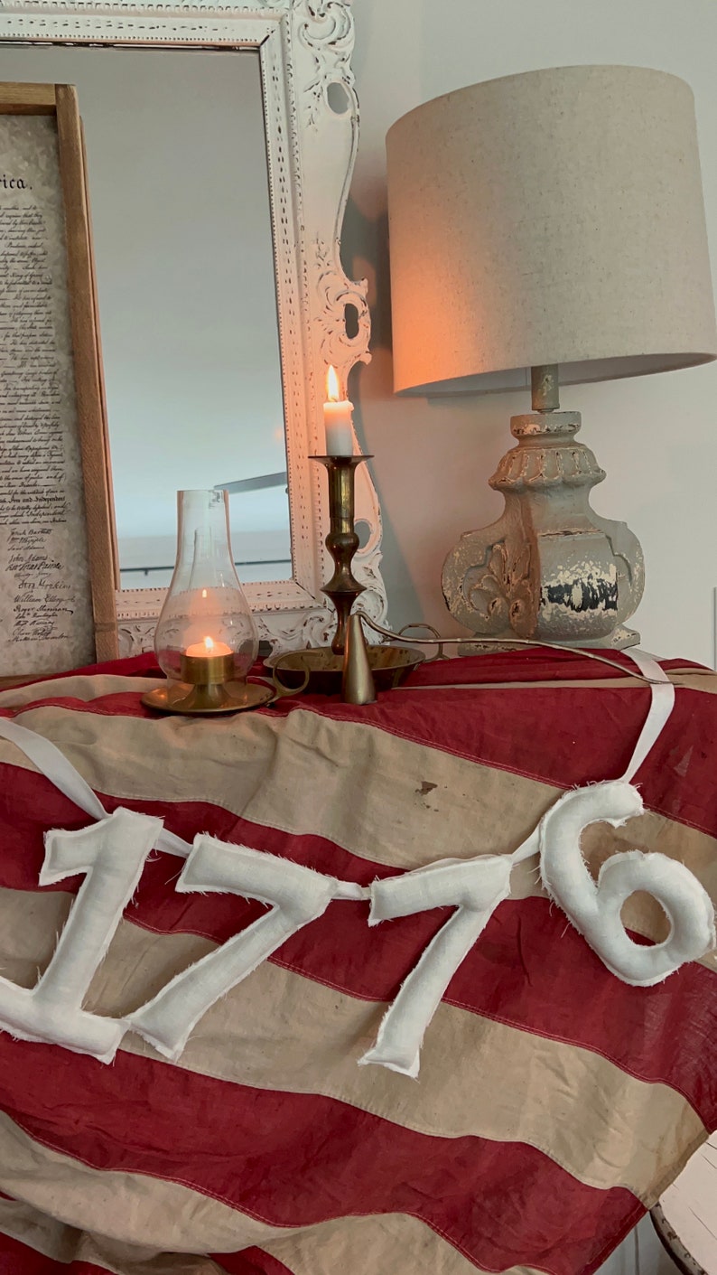 May include: A patriotic display featuring a vintage American flag with red and white stripes. The flag is adorned with a banner spelling out "1776". A lit candle sits in a brass holder, and a decorative lamp is visible.