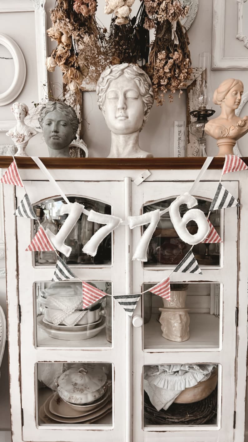 May include: A white cabinet with glass doors is decorated with a patriotic banner that reads "1776". The banner is made of white felt numbers and red, white, and blue striped pennants. The cabinet is filled with vintage dishware and busts.