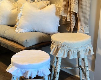May include: Two distressed white wooden stools with round seats covered in ruffled fabric. The stools have a rustic, vintage aesthetic, with peeling paint and a simple design. The background includes pillows, a bench, and decorative items.
