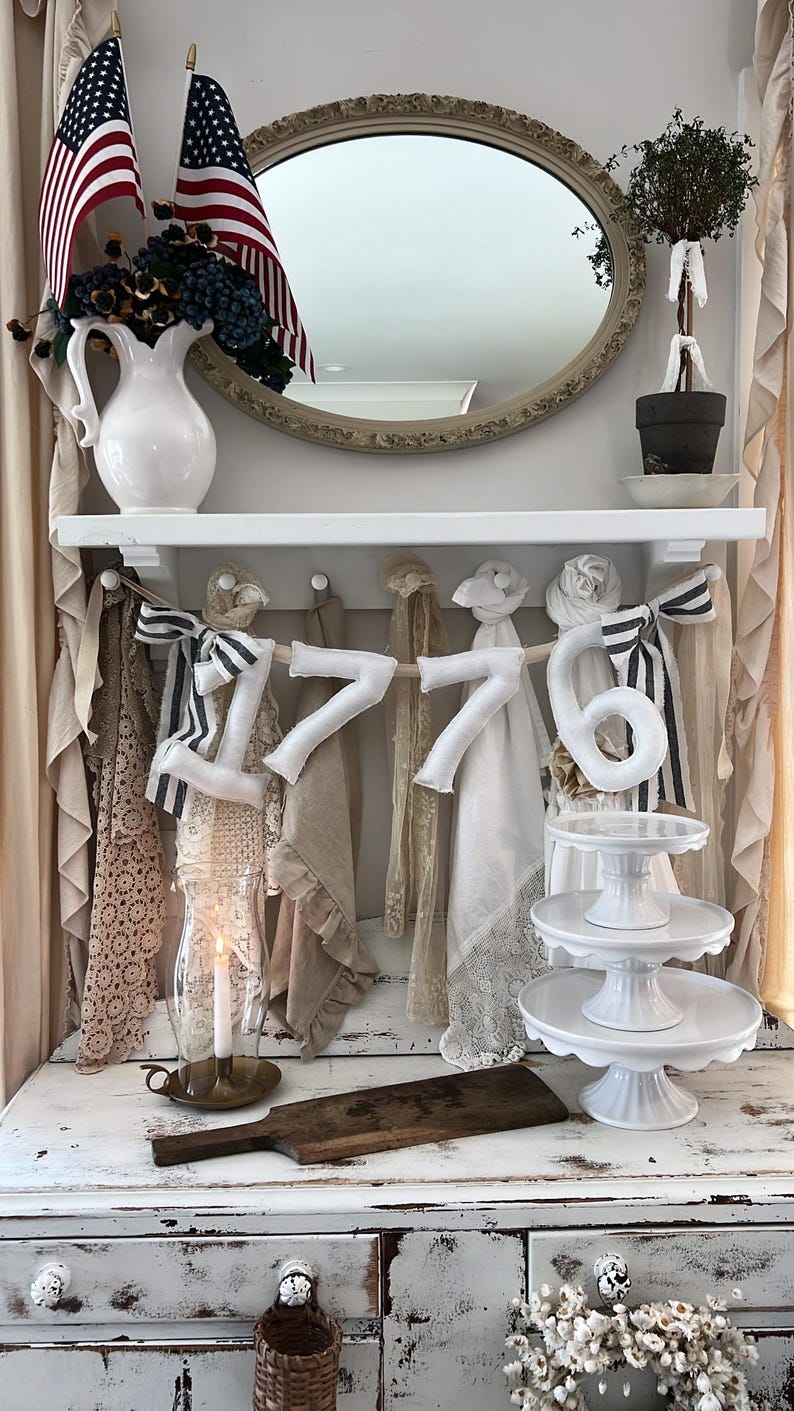 May include: A decorative display featuring American flags, a white pitcher with floral accents, and the number "1776" crafted from fabric. The display includes a tiered cake stand and a vintage-style wooden cutting board, evoking a patriotic theme.