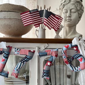 May include: A patriotic display featuring a decorative '76' banner made of fabric in red, white, and blue patterns, alongside miniature American flags in a white mug. A bust of a classical figure and a large, textured bowl are also visible.