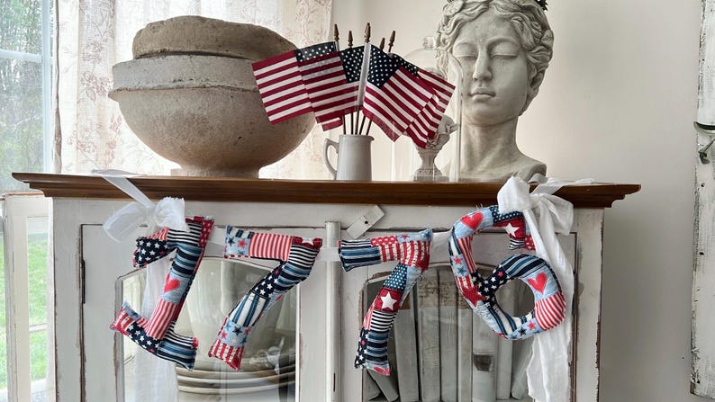 May include: A patriotic display featuring a decorative '76' banner made of fabric in red, white, and blue patterns, alongside miniature American flags in a white mug. A bust of a classical figure and a large, textured bowl are also visible.